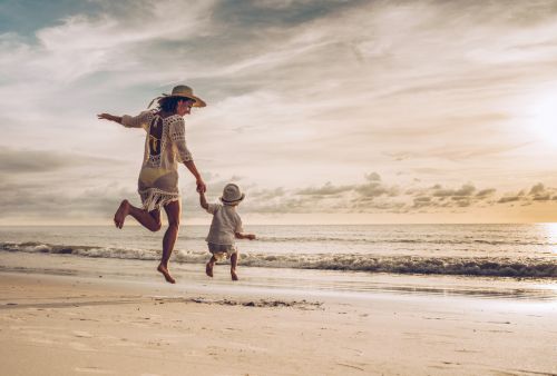 mother and son holding hands while jumping on the beach at sunset. - Ashley Wood Law, P.C.