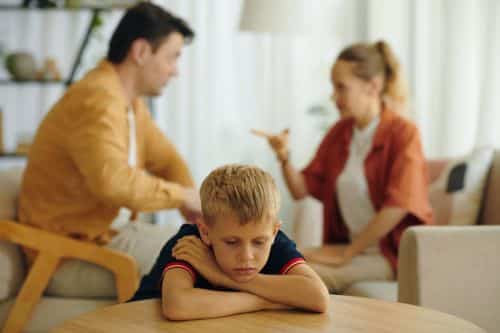 Sad child sitting at table and listening to quarrel of his parents in background