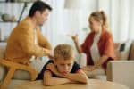Sad child sitting at table and listening to quarrel of his parents in background